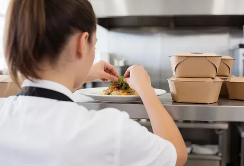 A chef in a white uniform and apron carefully garnishing a white plate with fresh ingredients in a bright stainless steel kitchen during active service