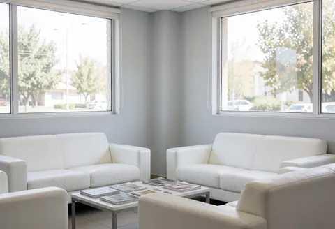 A clean modern waiting area featuring white sofas, a coffee table with magazines, and large windows letting in natural light for a professional office environment.