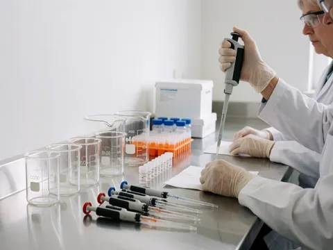 A medical professional in a clinical lab uses pipettes and a sample vial while wearing protective gloves and glasses on a shiny metal benchtop.