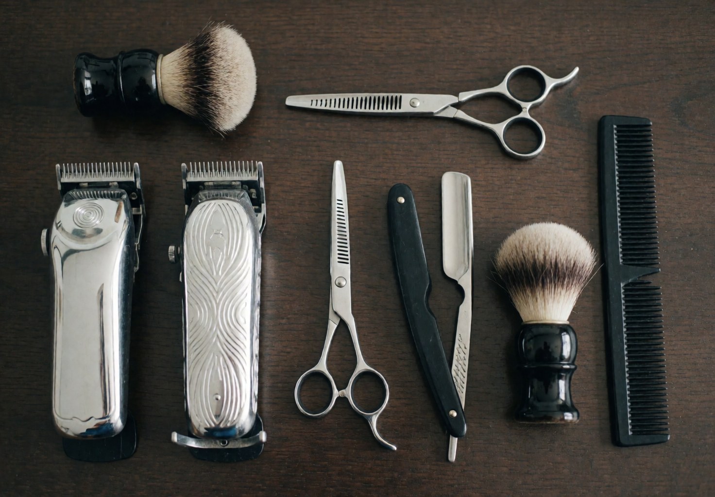 Top view of barber scissors, clippers, comb, and brush on a dark wooden surface with soft professional lighting