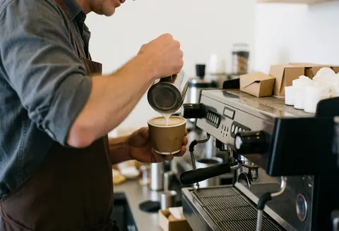 A close-up view of a barista preparing a latte with milk foam art in a paper cup at a coffee shop counter