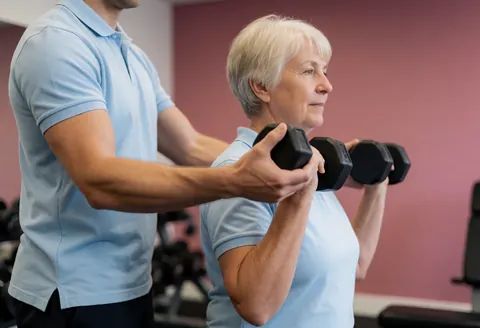 A fitness trainer helps an older client lift dumbbells during a gym session with natural light and a pink wall background.