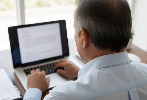 Back view of a man working on a laptop at a wooden desk with natural light, illustrating remote work or video calls