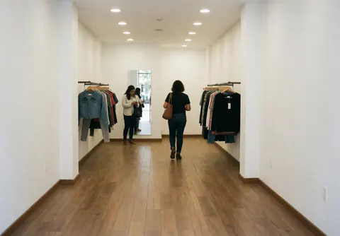 Interior of a modern clothing store featuring wooden floors and white walls, with two customers browsing garments on racks in a clean, well-lit space.