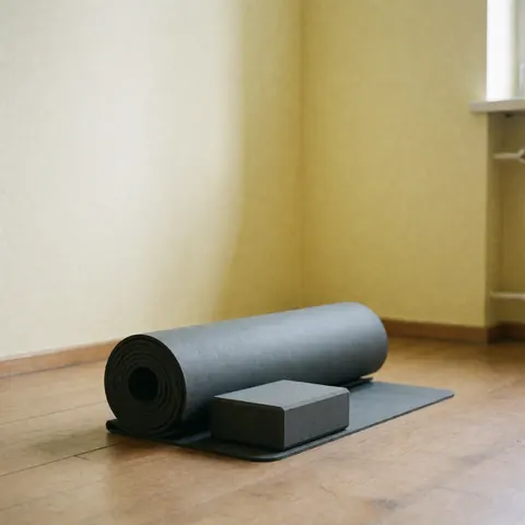 A rolled-up yoga mat and a rectangular yoga block resting on a wooden floor against a yellow wall, showcasing fitness equipment for wellness and meditation.