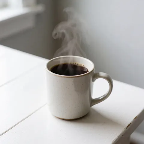 A white ceramic coffee cup sitting on a white surface with visible steam rising from the hot beverage, suitable for product display