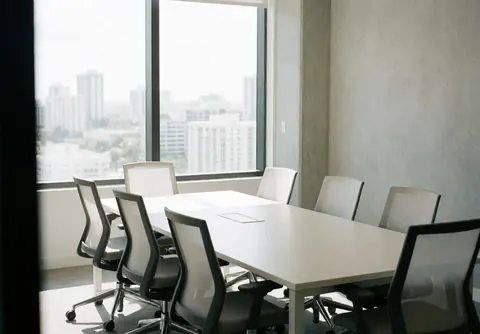 Modern conference room featuring a white table, ergonomic chairs, concrete walls, and a large window with a city view.