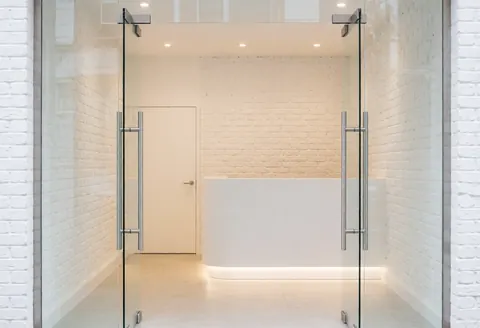 Modern gym entrance featuring glass doors, white brick walls, and a minimalist reception area with warm lighting and a clean professional atmosphere