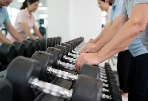 A row of dumbbells in a gym with adult hands selecting weights, featuring a mirror and equipment background with professional lighting