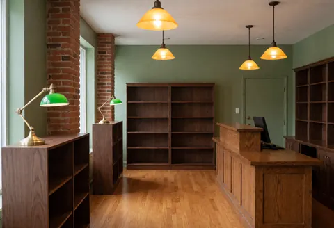 Empty bookstore interior featuring green walls, hardwood floors, walnut shelves, mahogany counter, hanging lights, and vintage reading lamps.