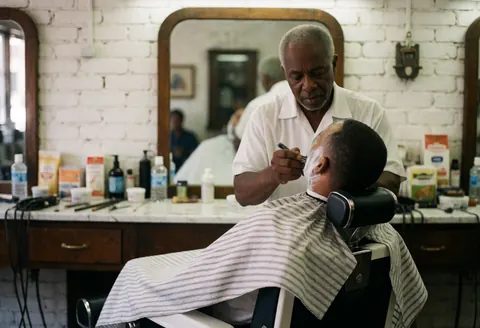 A senior barber carefully shaves a client's face while the customer sits in a barber chair inside a shop with brick walls and mirrors