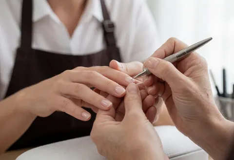 Close-up of a manicurist applying a nail treatment to a client's nails using a brush, with a blurred background and soft lighting.