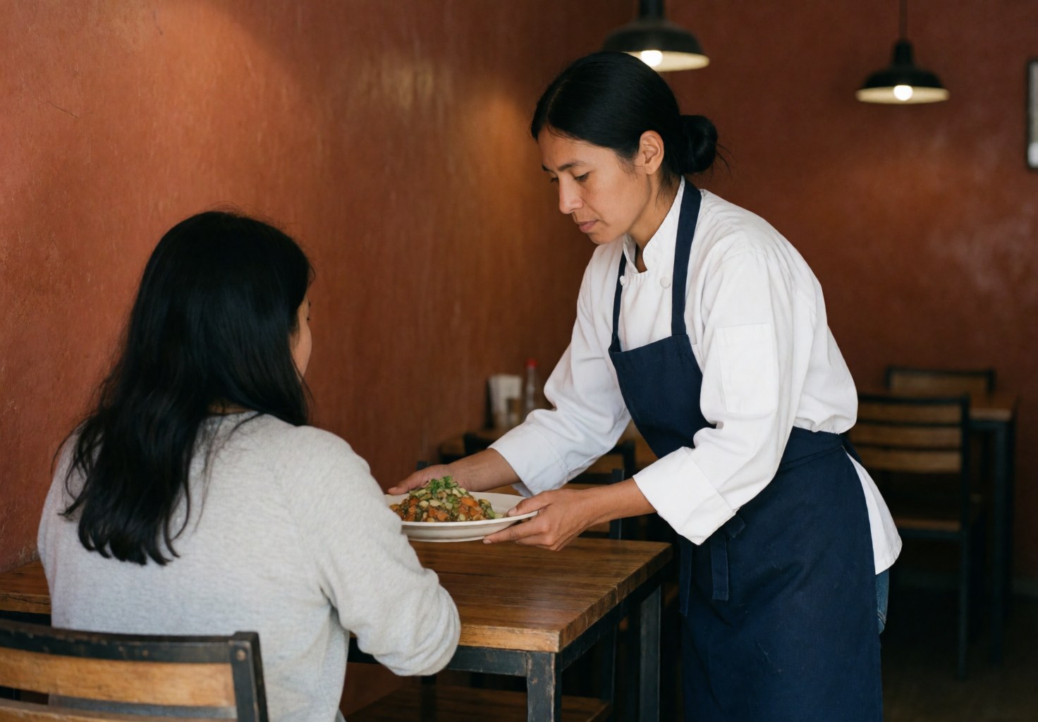 A cook serves a dish to a customer seated at a wooden table in a restaurant with terracotta walls and hanging lights.