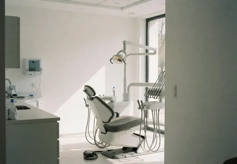 Bright modern dental office interior featuring a treatment chair, surgical light, and white walls illuminated by natural light from a window.