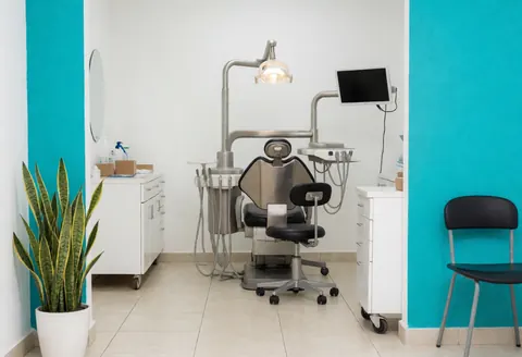 Interior of a modern dental clinic featuring a turquoise wall, white furniture, a black examination chair, plants, and bright clinical lighting.