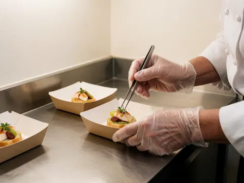 Close-up of a chef wearing latex gloves using tweezers to assemble a sandwich in a stainless steel kitchen.