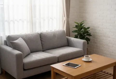 Cozy living room featuring a gray sofa, wooden table with tablet and coffee, white brick wall, curtains, and plant with natural light for studying or working from home