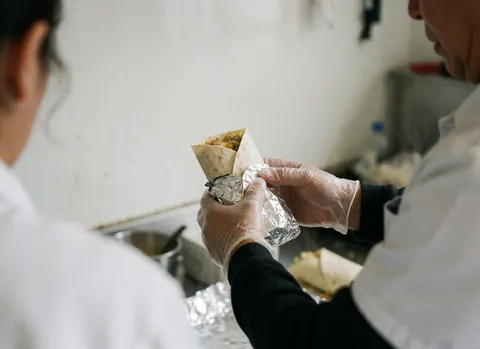Hands wearing gloves wrap a hot breakfast burrito in aluminum foil, with steam rising from the food truck kitchen interior