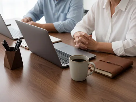 Two people working at a wooden desk with a laptop, coffee cup, notebook, and pen holder in a modern coworking environment