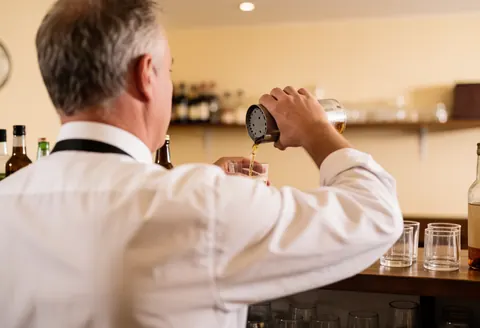 Bartender pouring a beverage into a glass behind a bar counter with warm lighting and a blurred beige background