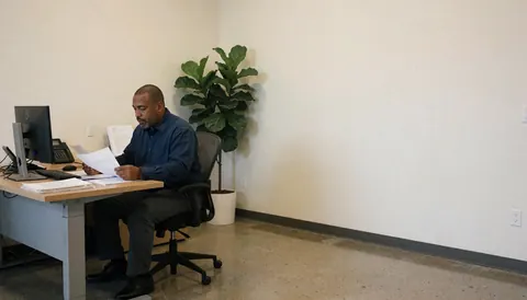 A man in a professional office setting reviewing paperwork at a desk with a computer, phone, and large plant in the background.