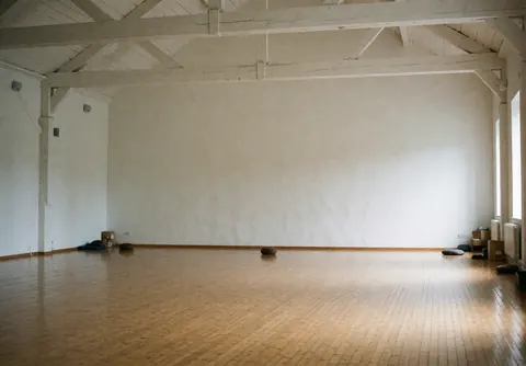 Empty yoga room featuring polished wood floors, exposed wooden beams, and white walls, creating a serene space for practice and meditation.