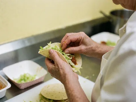 Close-up of hands assembling tacos with lettuce and sauce in a kitchen with yellow walls, showing ingredient texture and metal shine