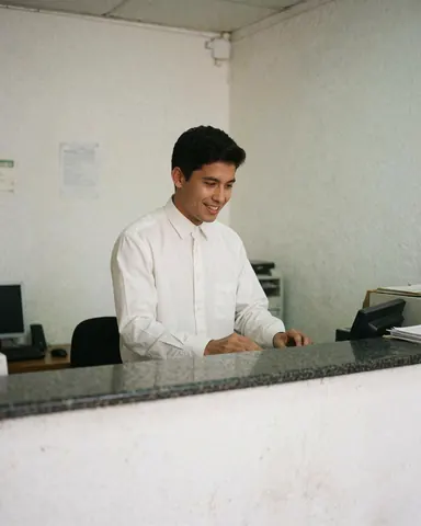 A smiling young bank employee in a shirt and tie stands behind a counter, focused on work in a professional white office environment.