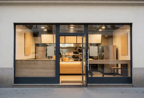 Modern fast food restaurant exterior featuring an open glass door, bright interior lighting, and a staff member working behind the counter.
