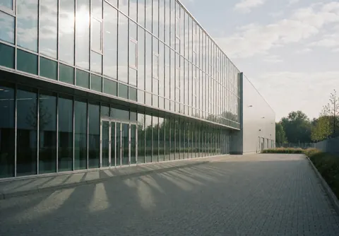 Exterior view of a modern logistics warehouse with large glass windows reflecting the sky and clouds, situated on a paved street in Hamburg.