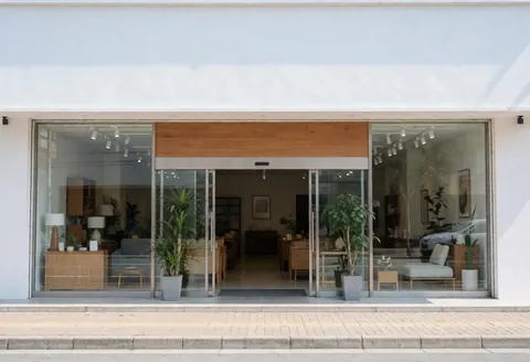 Clean white facade of a modern furniture shop featuring large glass doors, potted plants, and a paved sidewalk entrance.