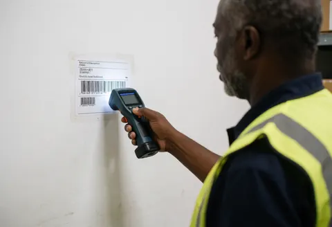 A worker in a safety vest scans a barcode on a white warehouse wall, demonstrating precise inventory management and logistics technology in action.
