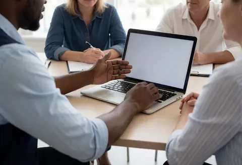 A professional meeting where a business proposal is being presented on a laptop to a client at a modern office desk.