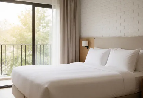 Spacious hotel room featuring a large bed, white brick wall, and window overlooking a balcony with natural light. Clean, minimalist, and inviting interior.