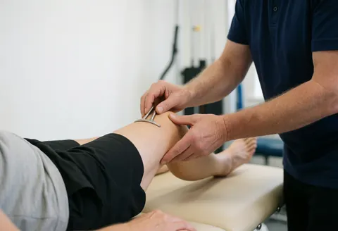 Physical therapist using a medical tool to treat a patient's knee on a treatment bed in a clean clinical setting with natural light