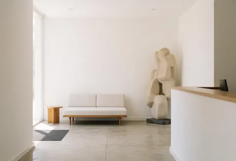 Modern minimalist reception area featuring an abstract sculpture, sofa, and natural light in a clean architectural space with concrete and wood elements.