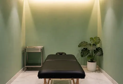 Minimalist massage room featuring green walls, white marble flooring, a black massage table, a monstera plant, and warm ambient lighting.