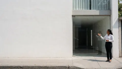 A professional female real estate agent stands outside a modern glass building entrance on a sunny day, gesturing with her hands while wearing business attire.