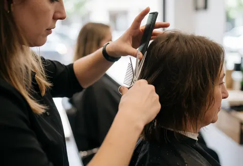 Professional hair stylist using scissors and comb to cut a seated client's hair in a salon with natural light and blurred background