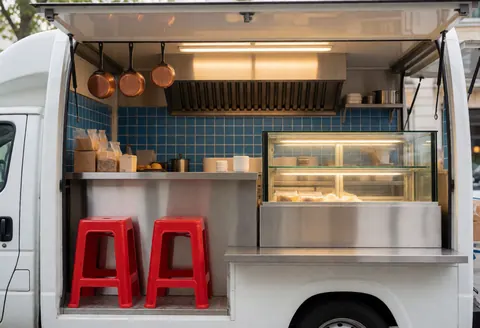 Spacious food truck interior featuring a stainless steel counter, blue walls, red chairs, and a display case with food under warm lighting.