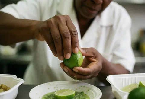 Close-up of a chef's hands squeezing a lime over a plate of green sauce, with a blurred street food background.