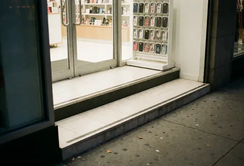 Clean and well-lit accessible storefront entrance featuring a glass door, step, and display cases for electronics and phones, welcoming customers to the business.