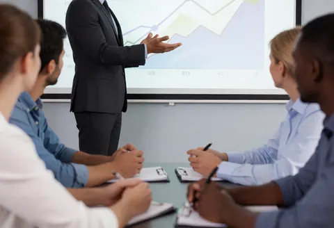 A professional presents growth charts on a screen to an attentive team seated at a table, taking notes with notebooks and pens.