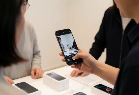 A salesperson holds a black smartphone to show the camera interface to a customer, with phone boxes on the table nearby