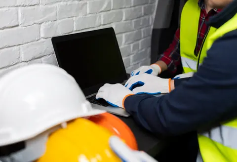 Construction inspector wearing helmet and safety vest working with a laptop near a white brick wall, wearing white gloves during a field inspection.