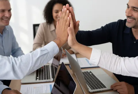 Three colleagues give each other a high five at a desk with laptops and documents during a professional office meeting.