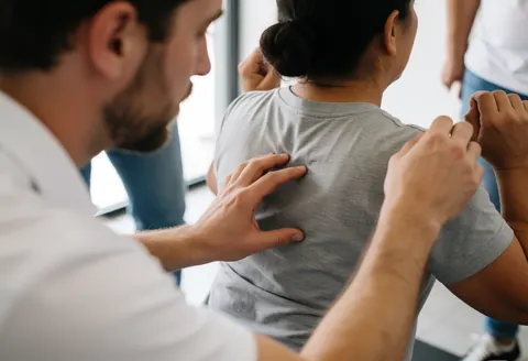 A fitness coach stands behind a client, using his hands to adjust and correct their spinal alignment and shoulder position during a strength training session.