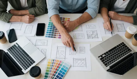 Design team working on web sketches at a desk with computers, phones, coffee, and color swatches