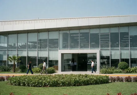 Modern industrial facility featuring a glass facade, green garden, and people walking near the main entrance under a clear blue sky.