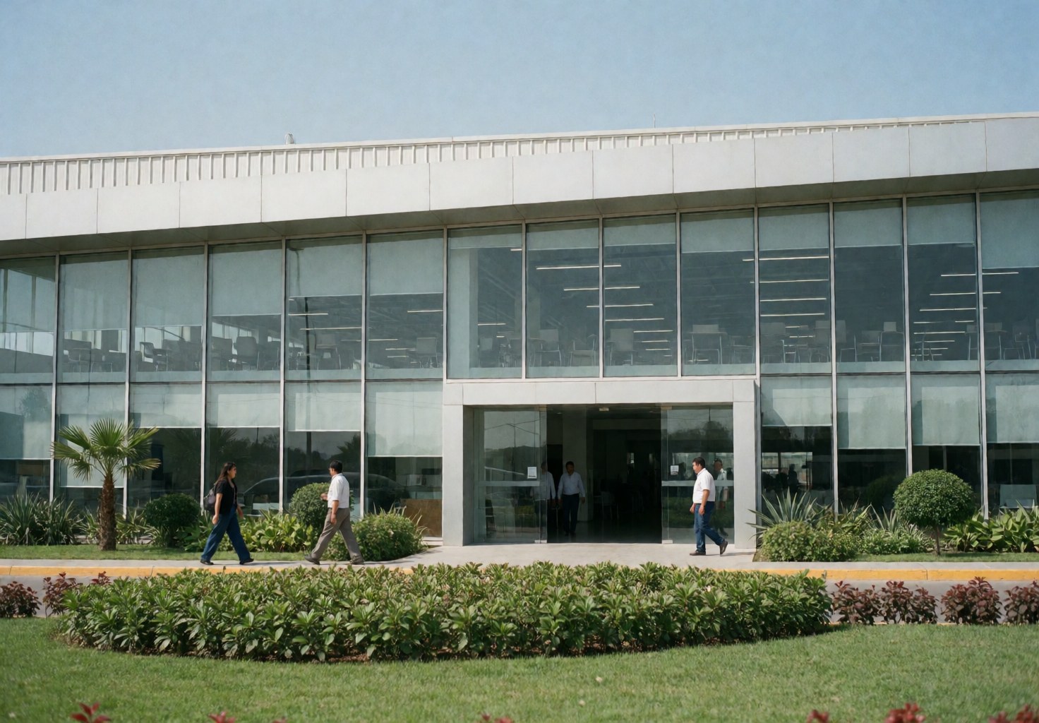 Modern industrial facility featuring a glass facade, green garden, and people walking near the main entrance under a clear blue sky.